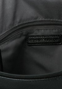 Zwarte binnenkant van een tas met een ritsvak en een leren label met textuur waarop "TOM TAILOR" is geperst. Gladde, synthetische voering zichtbaar.