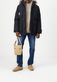 Svart isolerad jacka med pälsbesatt huva, tan sweatshirt, blå jeans och ljusa bruna skor. Håller en beige axelväska.