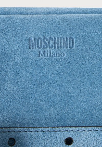 Gros plan sur un matériau en daim bleu embossé avec "Moschino Milano" et bord cousu avec deux trous ronds.