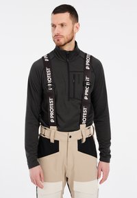 Heren outdooroutfit met een getailleerde zwarte top en beige sneeuwbroeken met zwarte accenten. Verstelbare banden met "PROTEST"-branding.