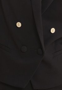 Blazer doppio petto nero con due bottoni logo dorati e due bottoni neri rotondi, dotato di tasche laterali e finitura testurizzata.