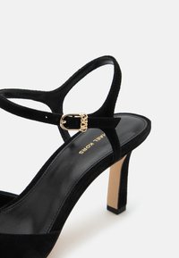 Sandale à talon haut en daim noir avec boucle dorée et talon fin, présentée sur fond blanc.