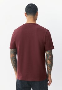 Burgundy short-sleeve T-shirt in a smooth fabric with a classic crew neck. Features a plain design and a relaxed fit, with detailed tattoos visible on the arms.