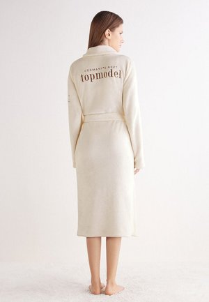 Woman standing barefoot wearing a cream-colored bathrobe with "Germany's Next topmodel" text on the back, looking sideways against a white background.