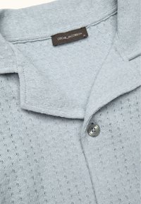 Lichtblauwe gebreide polotrui met een gebreid patroon, knoopsluiting en een kraag. Label toont "OSCAR JACOBSON" en maatlabel.