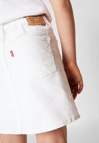 Jupe en denim blanc avec poches arrière, présentant la couture signature de Levi's et un patch en cuir camel sur la ceinture. Texture lisse.