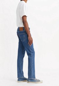 Blå denimjeans med rak benstil, med en brun läderlapp på baksidan, tillsammans med en vit t-shirt och blå sneakers.