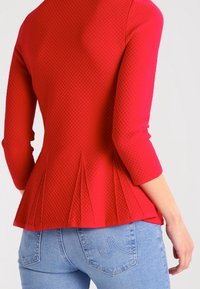 Haut texturé rouge à manches trois-quarts et design peplum, avec un motif géométrique. Associé à un jean bleu clair.