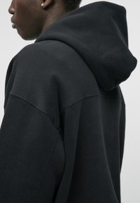 Musta kapuutsiga sviiter, mis on valmistatud pehmest kangast, suured kapuutsid, langetatud õlad ja nähtavad õmbluse detailid.