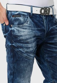 Denim jeans in een vervaagd donkerblauw met subtiele wassing. Bevat een witte riem met een metalen gesp en schuine naaddetails.