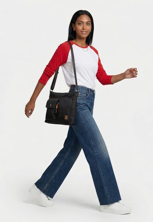 Femme marchant portant une chemise rouge et blanche à manches longues, un jean large bleu, des baskets blanches et un sac à bandoulière noir avec des poches avant.