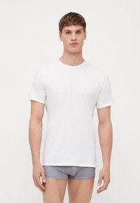 T-shirt en coton blanc avec un col rond et des manches courtes. Le tissu semble lisse avec un léger éclat, porté avec un boxer gris à rayures.