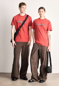 Camisetas de algodón rojo con el logo "tommy jeans" combinadas con pantalones anchos marrones. Una modelo lleva un bolso negro de hombro, ambas usan zapatos negros.