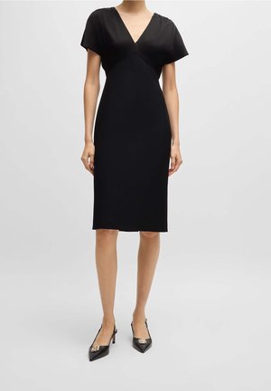 Mujer que lleva un vestido negro hasta la rodilla con mangas cortas y escote en V, combinado con tacones puntiagudos negros con adornos metálicos.