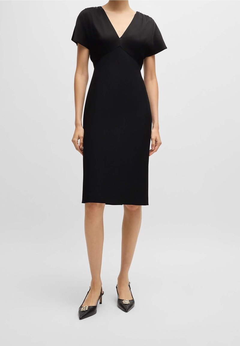 Femme portant une robe noire mi-longue à manches courtes et col en V, associée à des escarpins noirs à bout pointu avec des ornements métalliques.