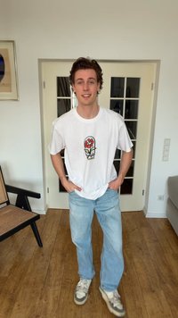 Νεαρός άνδρας που στέκεται σε εσωτερικό χώρο, φοράει λευκό μπλουζάκι με κόκκινο σχέδιο λουλουδιού, ανοιχτόχρωμο τζιν και λευκά αθλητικά, με τα χέρια στις τσέπες.