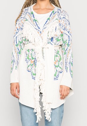 Mujer con cárdigan blanco de punto con patrones multicolores, flecos y un cinturón atado sobre vaqueros azul claro.