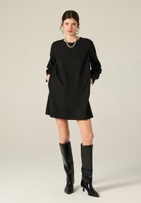 Donna che indossa un mini abito nero a maniche lunghe, collana in argento, orecchini a cerchio e stivali neri al ginocchio con tacco, in piedi con le mani nelle tasche.