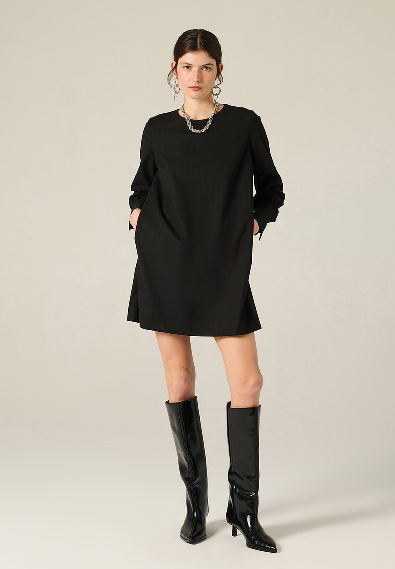 Donna che indossa un mini abito nero a maniche lunghe, collana in argento, orecchini a cerchio e stivali neri al ginocchio con tacco, in piedi con le mani nelle tasche.