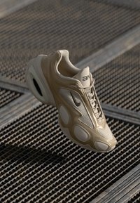 Shoe atlético bege com parte superior em malha texturizada, sobreposições suaves e grandes recortes ovais. Apresenta um colarinho acolchoado e fecho com atacadores.