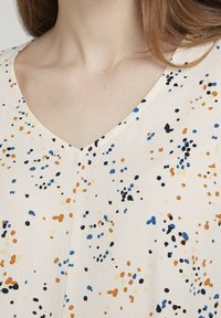 V-Ausschnitt-Bluse aus leichtem Material mit einem cremefarbenen Hintergrund, der mit verstreuten blauen, orangefarbenen und schwarzen Sprenkeln verziert ist. Glatte, weiche Textur.
