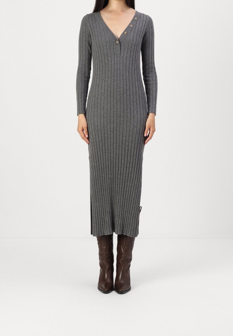 Femme portant une longue robe tricotée gris côtelé avec un col en V et trois boutons, associée à des bottes en cuir marron mi-mollet, debout sur un fond blanc.
