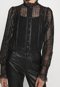 Blouse en dentelle noire avec motifs floraux complexes, silhouette structurée, fermeture à boutons, et col ainsi que poignets à volants.