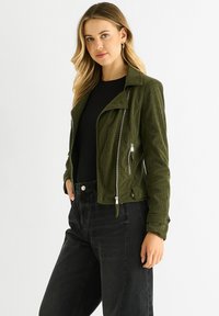 Olive green corduroy jacket with a collar, silver zippers, and a cropped fit, worn over a black top with black high-waisted jeans.