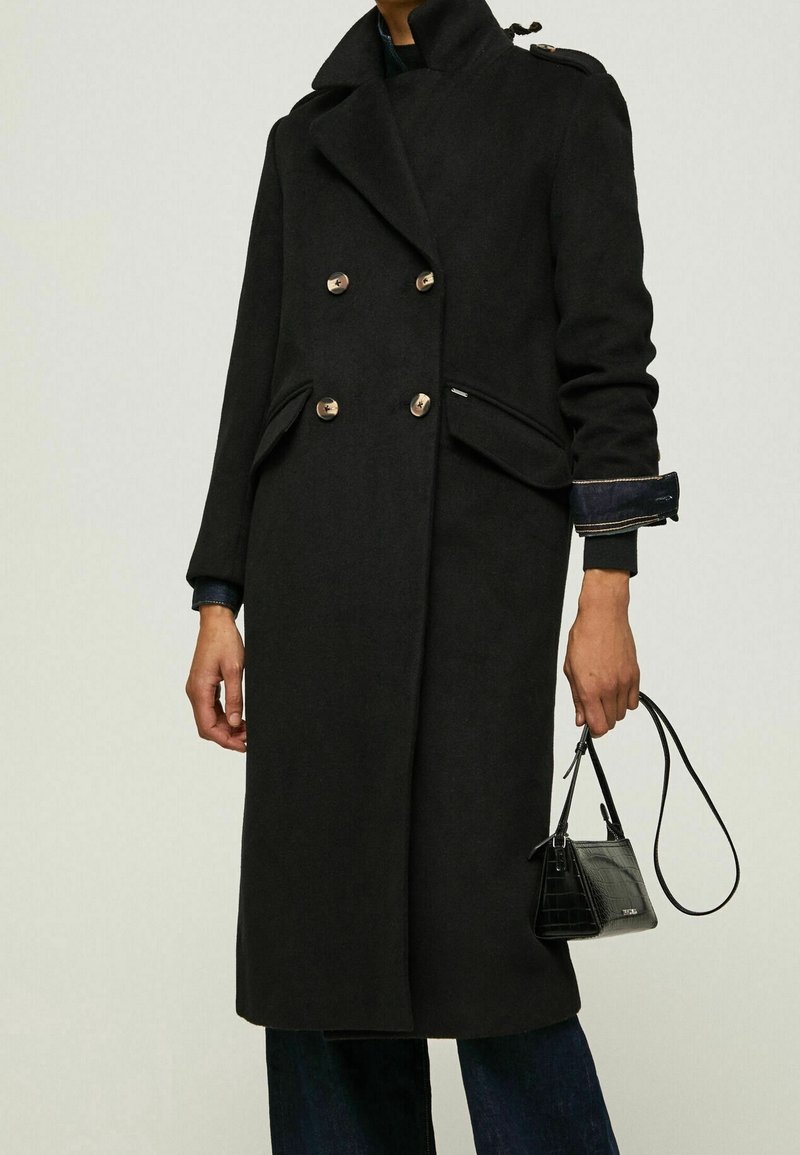 Personne portant un long manteau noir à double boutonnage avec des boutons dorés, tenant un petit sac à main noir avec une fine bandoulière.