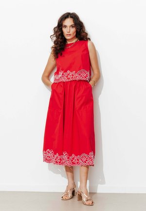 Femme aux cheveux bouclés portant une robe rouge sans manches avec une broderie florale blanche, debout contre un mur blanc, mains dans les poches.