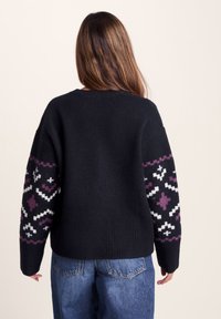 Woman with long brown hair wearing a black knit sweater with purple and white patterned sleeves and blue jeans, facing away from the camera.