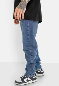 Blå denimjeans med rakt ben som har blekna områden och kontrasterande sömmar, tillsammans med svarta och vita sneakers.