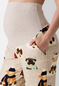 Schwangere Person trägt beige Umstandshose mit hohem Bund und Winterkuhprints, Hand in der Seitentasche.