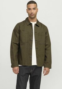 Giacca in tela verde oliva con collo a camicia, chiusura a bottoni, due tasche sul petto e maniche lunghe, indossata sopra una camicia bianca.