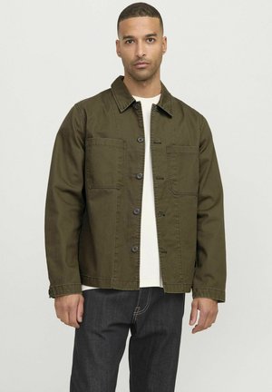 Olijfgroene canvasjack met een kraag, knoopsluiting, twee borstzakken en lange mouwen, gedragen over een wit shirt.