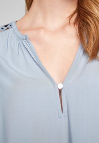 Vaaleansininen paita, joka on valmistettu pehmeästä ja kevyestä kankaasta. Ominaisuuksiin kuuluu V-muotoinen kaula-aukko, metallinen nappi ja yksityiskohtainen ompelu olkien kohdalla.