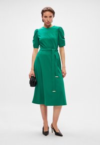 Πράσινο midi φόρεμα με κοντά φουστανάκια, εφαρμοστό μπούστο, ζώνη στη μέση και φουσκωτό σιφόν. Μαύρη τσάντα και μυτερές ψηλοτάκουνες γόβες ολοκληρώνουν την εμφάνιση.