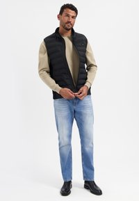 Schwarze gefütterte Weste mit hohem Kragen, getragen über einem beigen Pullover. Hellblaue Jeans und schwarze Stiefeletten runden das Outfit ab.