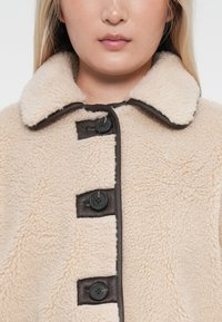Beige faux shearling coat with a dark brown collar and buttons. Features a soft texture and four black buttons down the front.