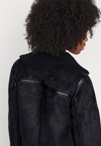Chaqueta de ante negra con un suave cuello de borrego, con detalles de cuero y un acabado texturizado; diseño ajustado con costuras visibles.