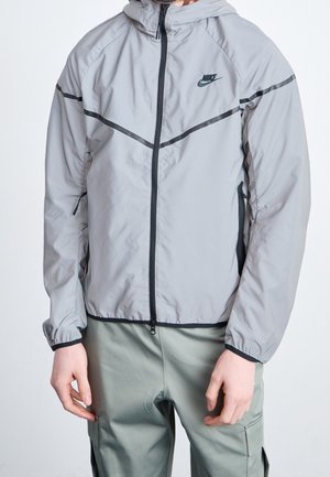 Persona vistiendo una chaqueta cortavientos con capucha Nike de color gris claro con cremallera negra y detalles en negro, combinada con pantalones cargo verdes.