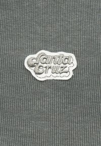 Γκρι ριμπ ύφασμα με ένα λευκό περιμετρικά πλαίσιο και κέντημα με το κείμενο "Santa Cruz" σε στυλιζαρισμένα πεζά γράμματα.