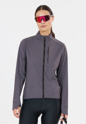 Femme portant une veste de cyclisme violet-gris, un short de cyclisme noir rembourré et des lunettes de soleil de sport rouges réfléchissantes, tenant une sangle d'accessoire noire.