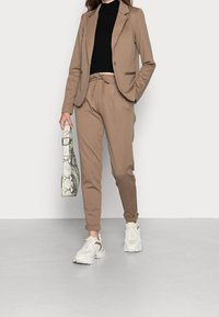 Blazer beige avec poches latérales sur un col roulé noir à côtes, associé à un jogging beige, des baskets blanches et un sac en toile à motif serpent.