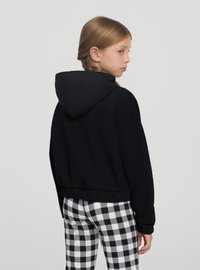 Felpa nera con cappuccio, dal design corto e orlo elastico, abbinata a pantaloni a quadretti bianchi e neri, su uno sfondo semplice.