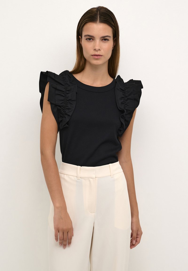 Haut noir avec manches courtes à volants, col rond et tissu texturé. Porté avec un pantalon large beige clair, au fini lisse.