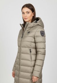Beige puffatakki hupullinen, vetoketjulla edessä ja sivutaskuilla. Sisältää tikkikuvion ja patchin vasemmassa hihassa.