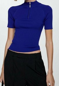 Prążkowany, niebieski top z krótkim rękawem, z wysokim kołnierzem zapinanym na zamek. Dopasowany krój i krótka długość. W połączeniu z czarnymi spodniami.