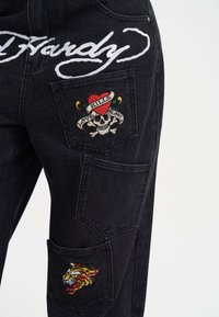 Pantalones de mezclilla negros con costuras de logotipo blanco prominentes, bordados de calavera y corazón, y un parche de tigre en el bolsillo lateral.