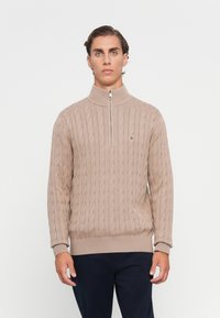 Man staat rechtop met het gezicht naar voren, gekleed in een beige kabelgebreide zip-up trui en donkere broek, voor een effen witte achtergrond.